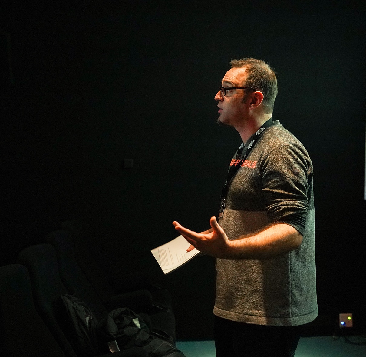 A man in a dark room, wearing a grey shirt and lanyard, appears to be giving a presentation.